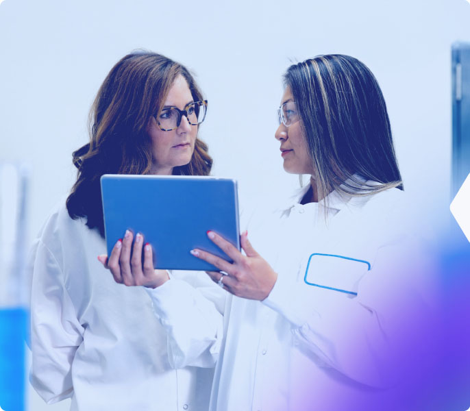 Two female scientists in lab coats reviewing data on a tablet in a laboratory setting.